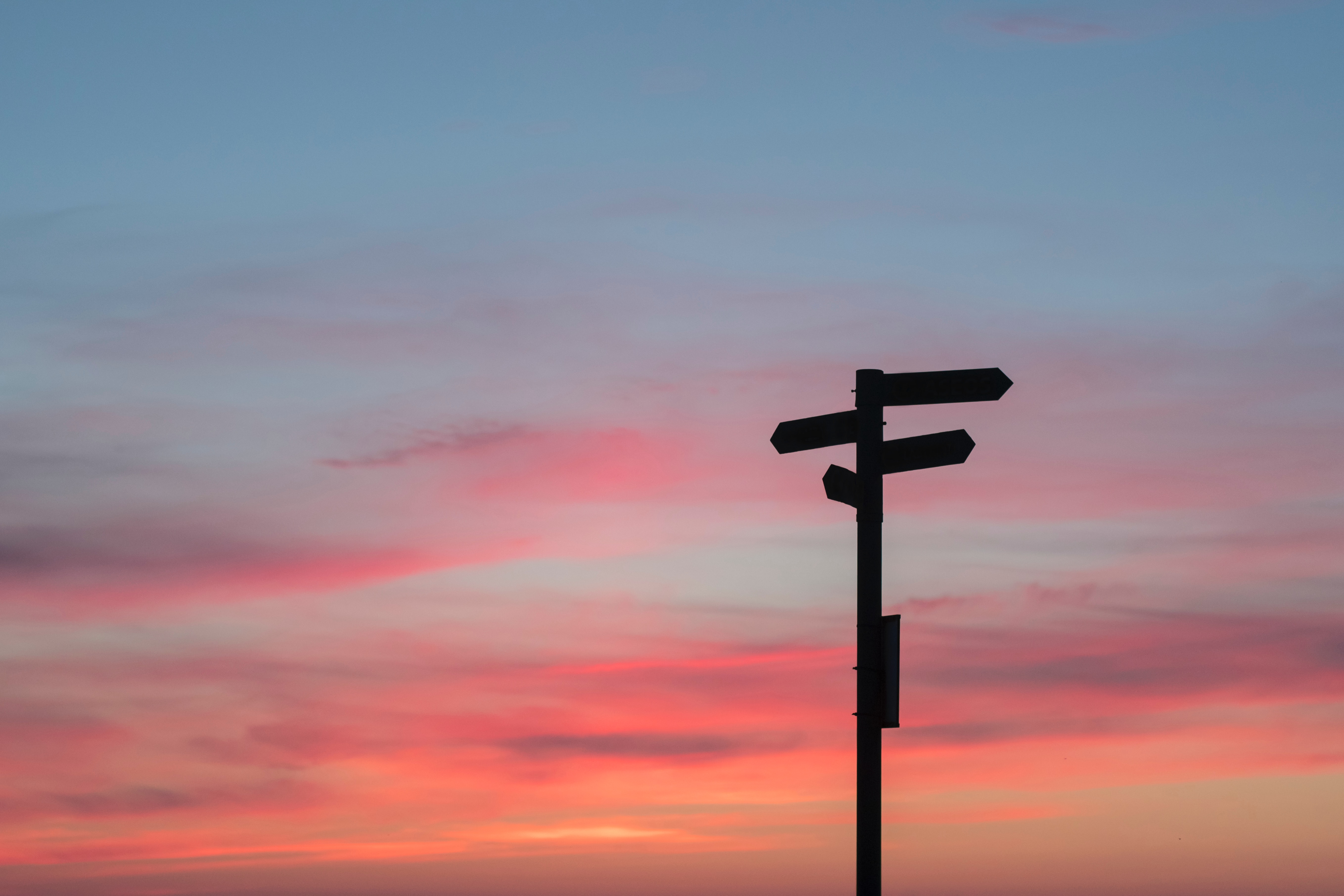 A signpost pointing in four different directions. It is in silhouette against a pink and grey sunset sky.