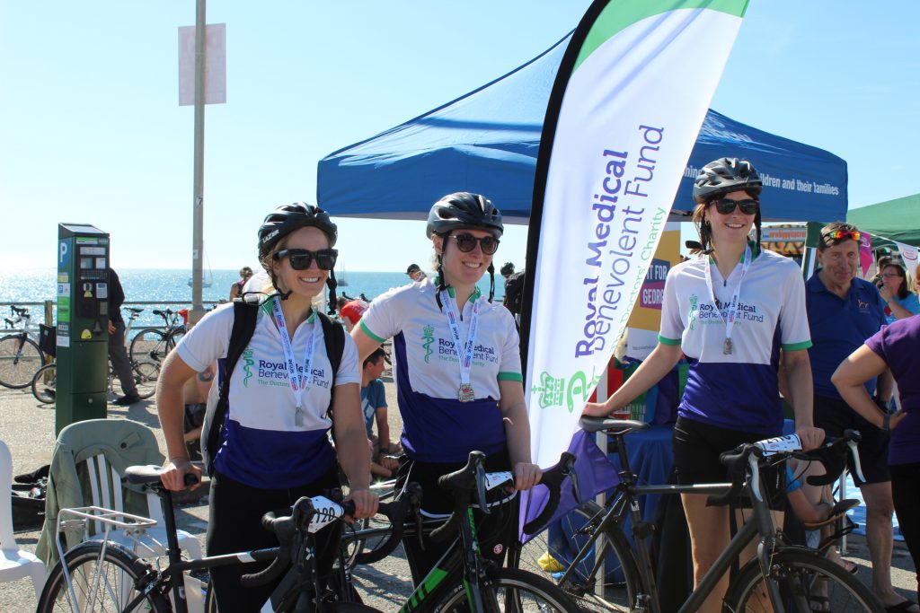 Three cyclists, Elizabeth Nally, Sarah Hancox, and Lizzie Kostev, standing with their bikes in the sun at the Brighton seafront, wearing RMBF cycling tops