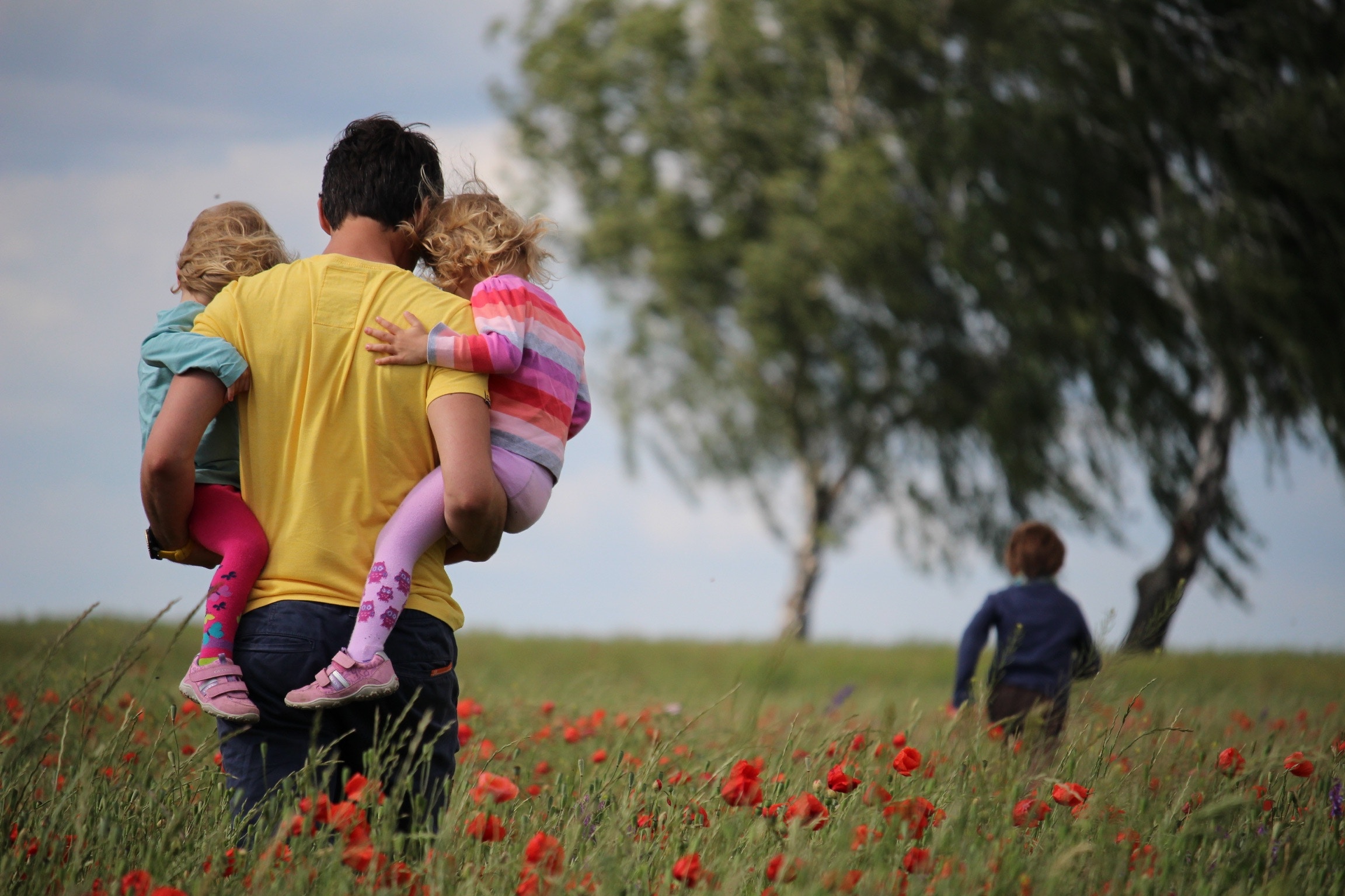 A view from behind of a man who is carrying two small children, one on each arm. They're walking through a field of flowers towards some trees. An older child runs ahead of them.