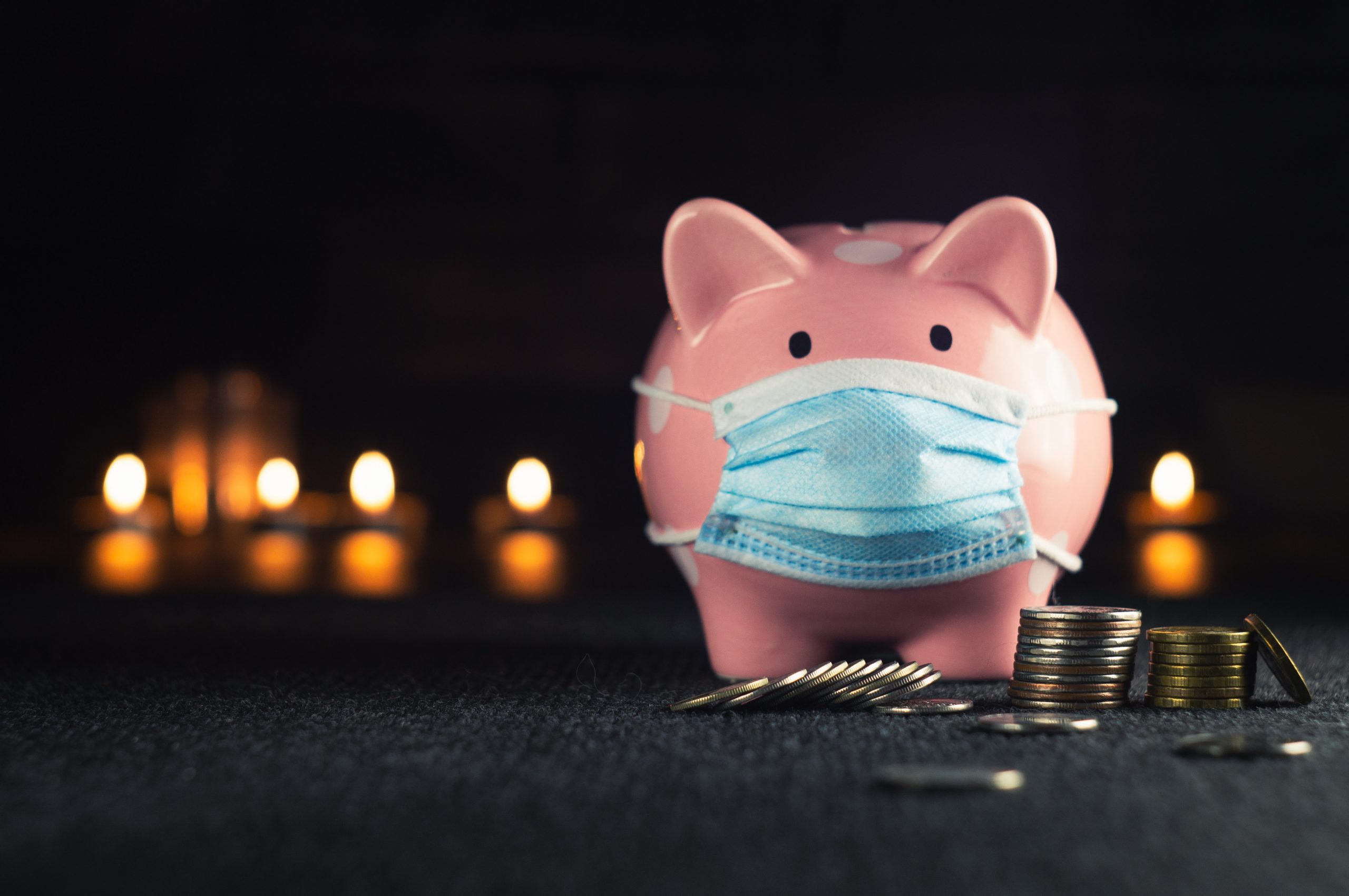 A piggy bank wearing a surgical mask. Coins in small piles are arranged in front of its feet.