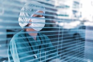 A doctor in scrubs and a face mask stares out through the slats of a window blind, with a faraway look. They seem lonely, worried, anxious.