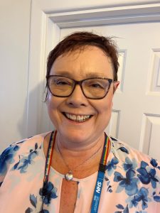 A picture of interviewee Dr Diana Cassell. She is a woman with short red-brown hair and glasses. She is smiling and wearing an NHS rainbow lanyard.
