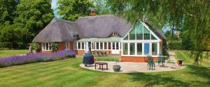 A picture of the cottage where Acaia Retreat is held. It has a thatched roof and a conservatory. Outside is a patio with furniture, a neatly mown law, and a patch of heather.