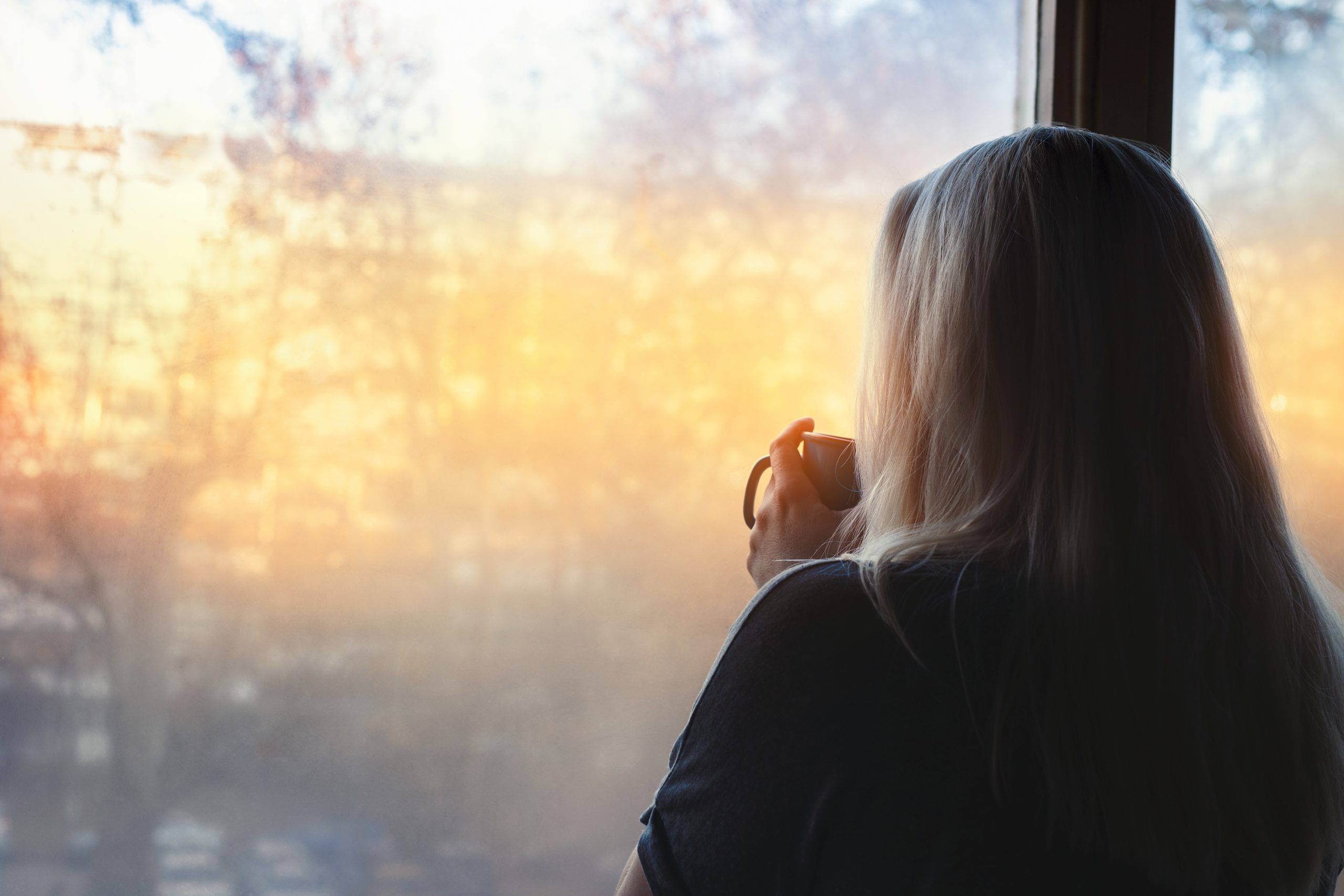 A woman with white hair looking out of a window at the sunrise. She is facing away from the camera, and holding a mug, close to her face, maybe her morning coffee.