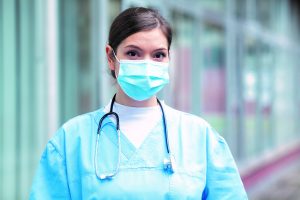 Photo of a young female doctor in scrubs, wearing a surgical mask. She looks calm and composed.