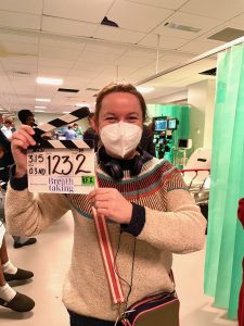 Dr Rachel Clarke on set. She is a white woman with dark hair. She is wearing a wooly jumper, a high quality face mask, and has headphones around her neck. She is holding a clapperboard.