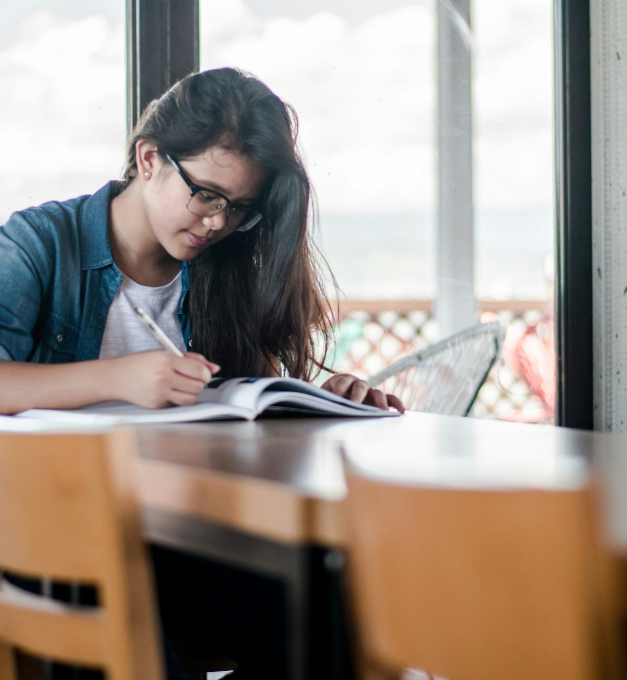 A light-skinned young woman with long dark hair sits at a table, studying from a textbook. She is wearing glasses, and a denim shirt over a t-shirt.