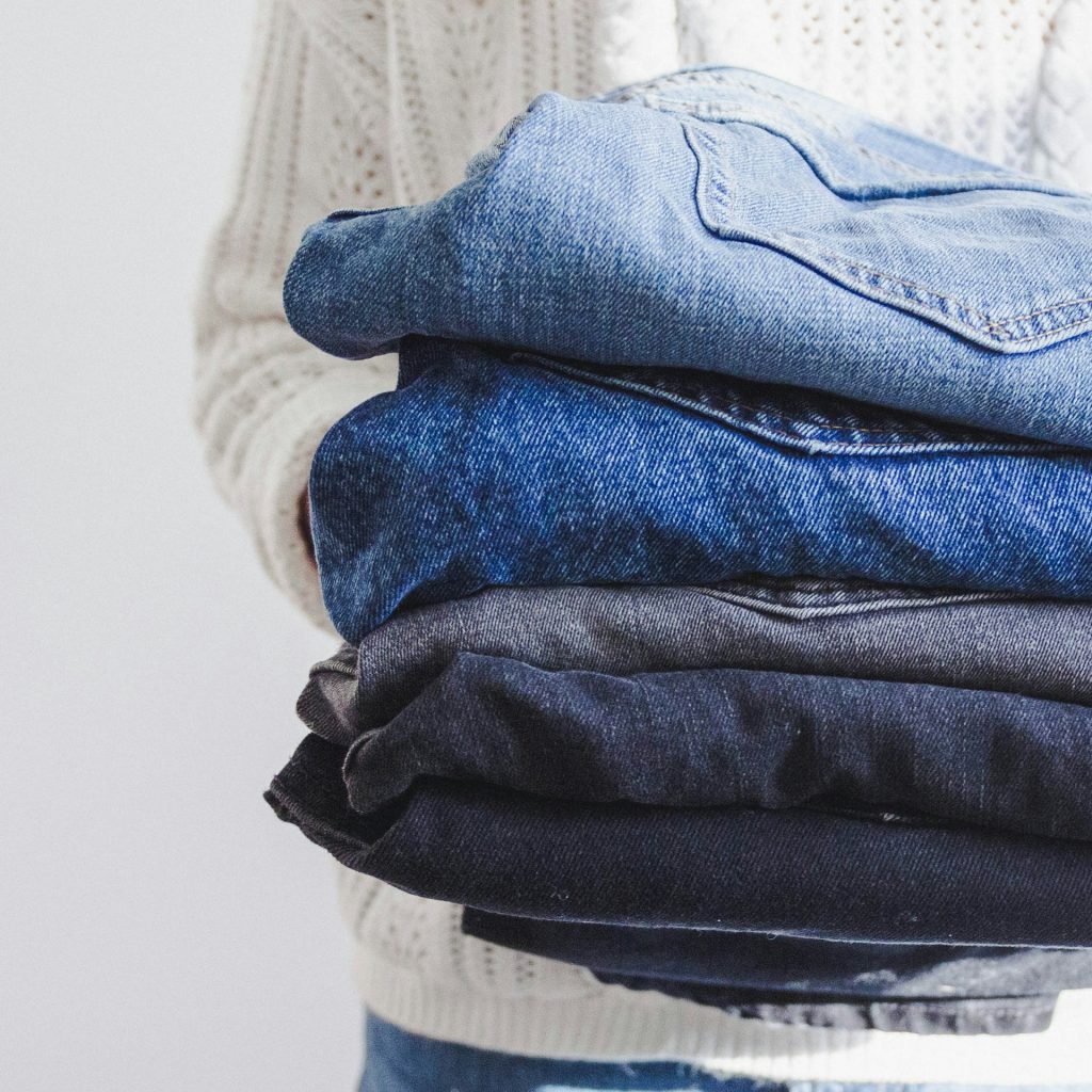 A person in a white jumper, holding 5 pairs of different blue shade jeans 