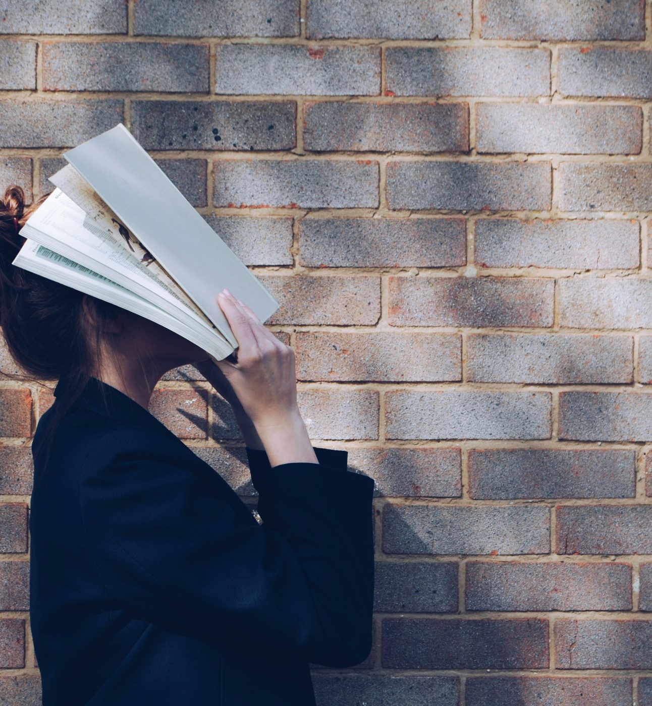 A young light-skinned woman stands outside a building. She is looking up and fully burying her face in a book, perhaps feeling a little overwhelmed.