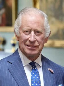 A head-and-shoulders photograph of His Majesty King Charles III, taken in 2023. He is an older white man with short grey hair. He is wearing a blue pinstripe suit with a small flower on the lapel, a white shirt, and a blue tie.