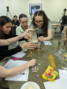 Three young women are trying to build a tower out of jelly babies and spaghetti. They're laughing, and it looks like the tower might fall if they let go of it.