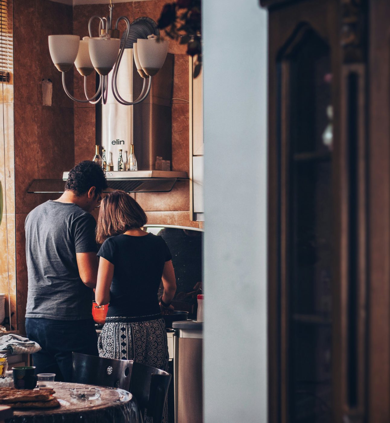 A photo of a couple cooking together in a rustic-looking kitchen. They are both facing away from the camera, concentrating on something on the stove. Soft afternoon light comes in from the windows.