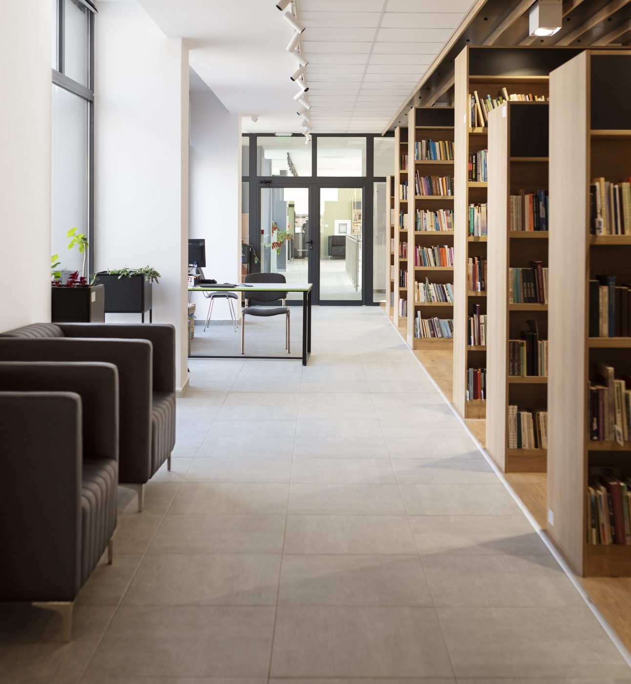 A photo of a corridor in a modern library building. We can just see into the aisles of bookshelves on the right. Light comes in from windows on the left, where there are also some leather chairs, plants, and an information desk.