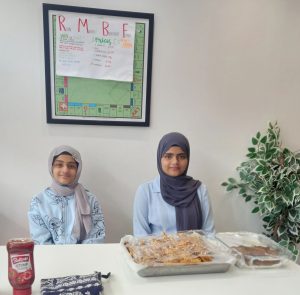 Nivisha and another young woman smiling, sat behind a table with food displayed on plates