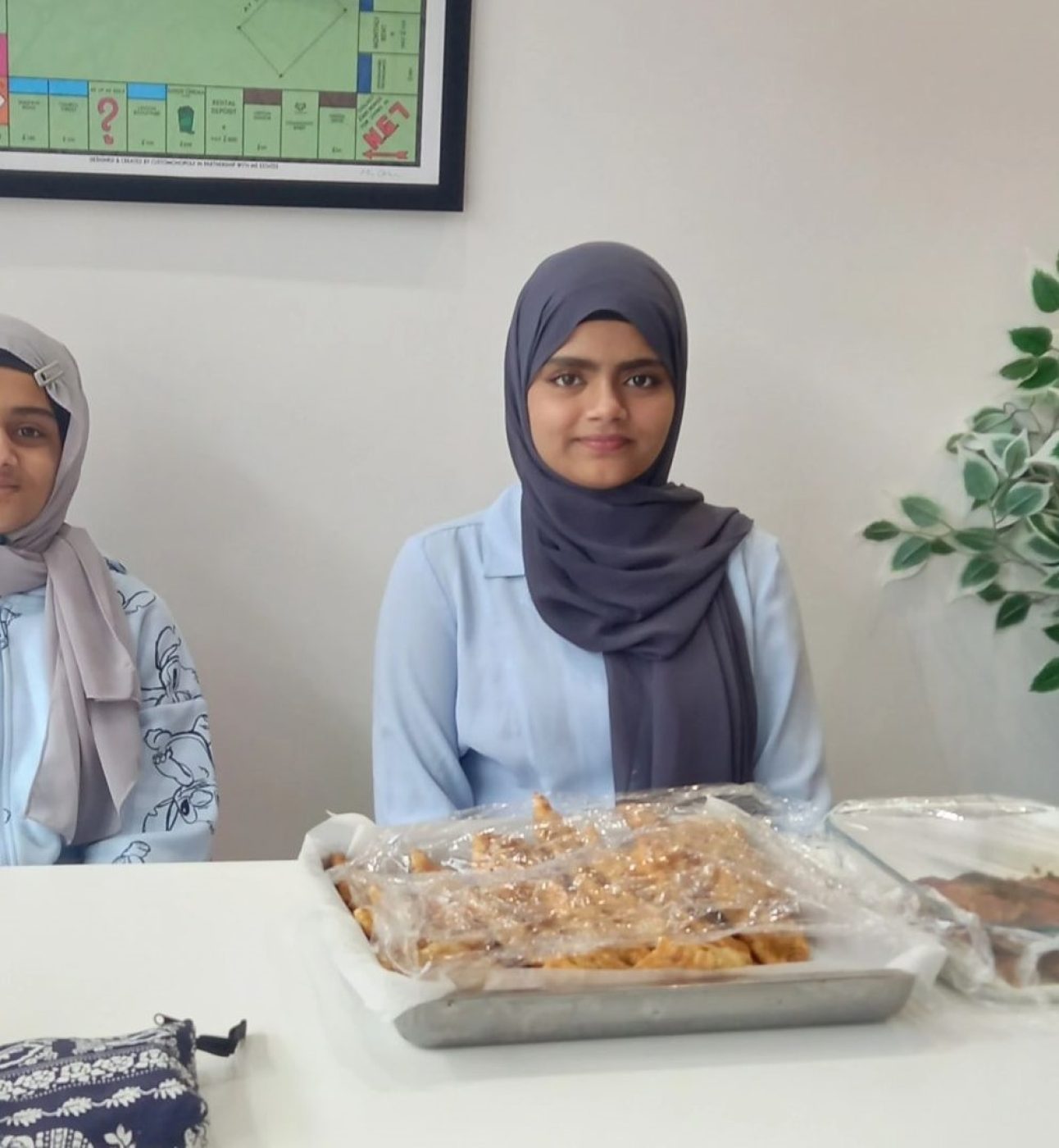 Sareena and another young woman smiling at the camera, sat behind a table with food displayed