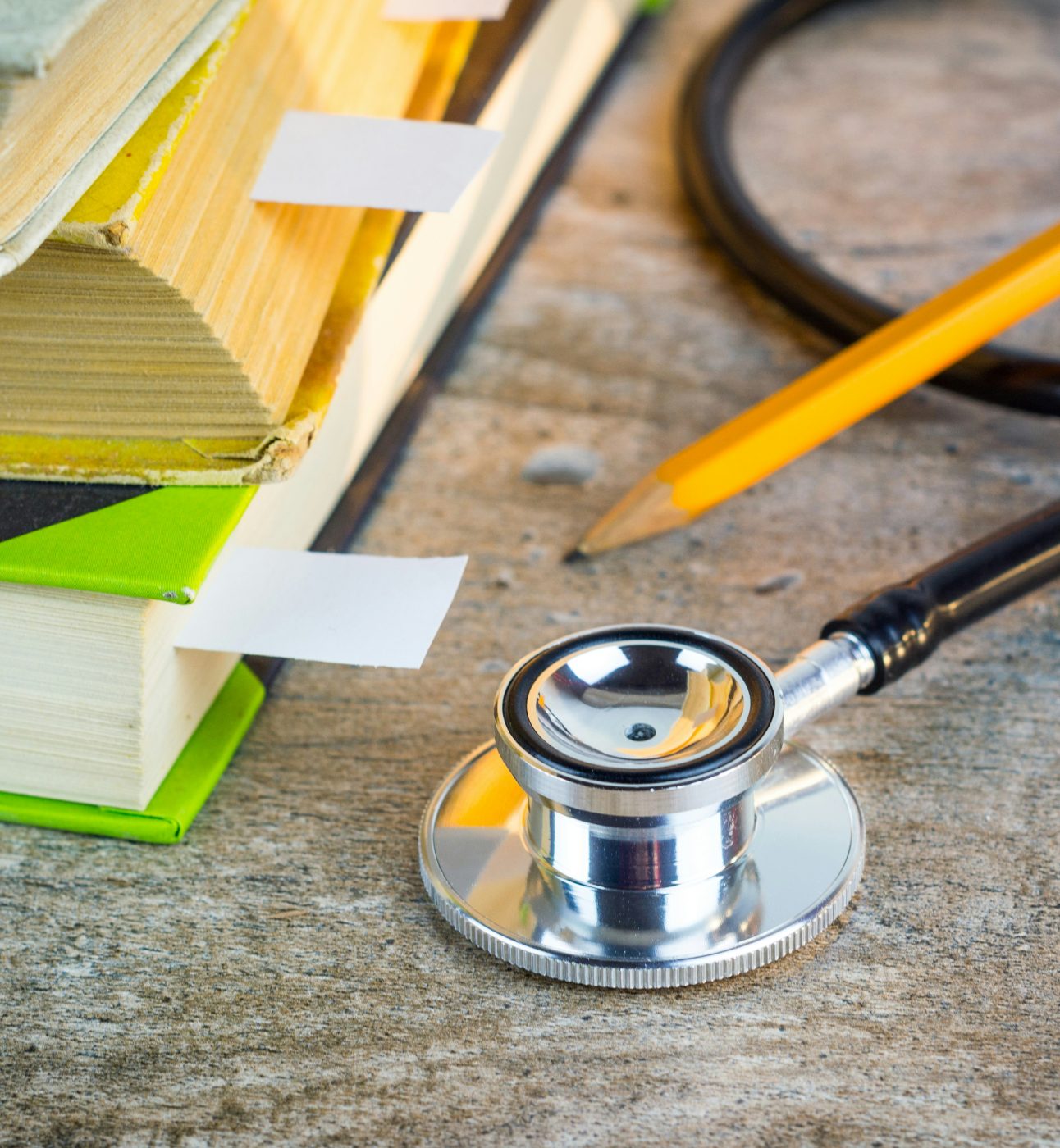 A close-up photograph of some objects on a table: the round end of a stethoscope, a yellow pencil, and some textbooks with tabs marking certain pages.