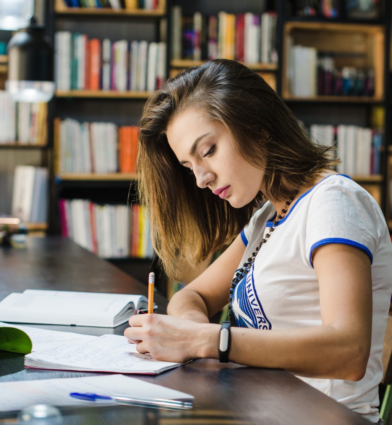 A young woman sits at a long desk in a library, writing in her notebook - she seems to be concentrating intently. She has white skin and shoulder-length brown hair. She's wearing a blue and white t-shirt and a bead necklace.