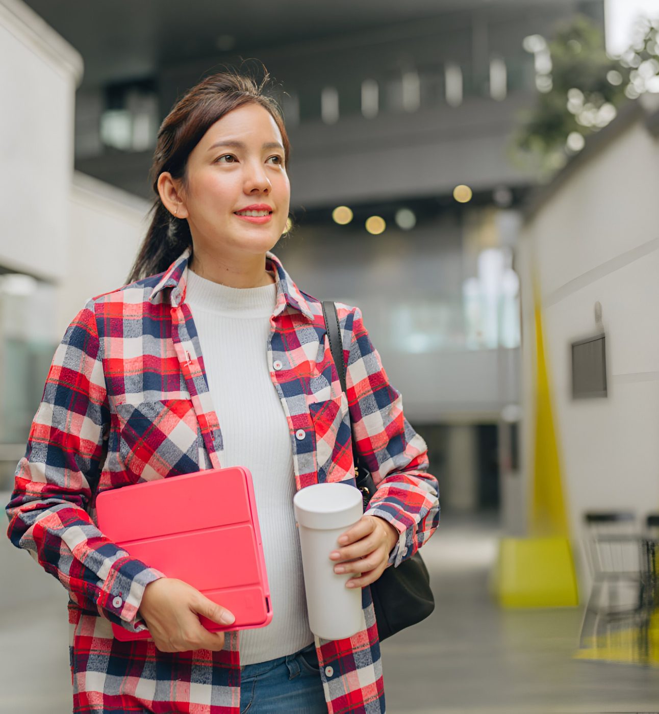 A young woman with pale skin and long dark hair walks down a university hallway, looking relaxed and confident. She's carrying a reuseable coffee cup and a pink tablet case.