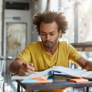 A young man, wearing a yellow t-shirt, with brown skin and mid-length afro hair, sits at a table in a well-lit room. He's studying, making pencil notes in an exercise book, looking deep in concentration.