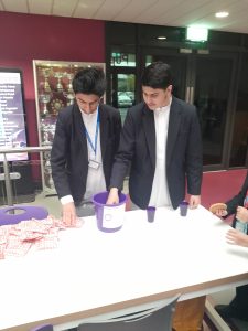 Two male students stood in front of a table, one checking the donations in a purple bucket, one student sorting red bags (of sweets).