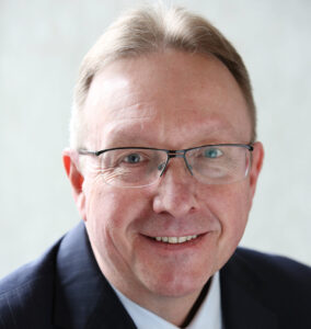A picture of RMBF CEO Steve Crone,. He is a white man in his 60s with light hair. He wears a suit with a collared shirt, and glasses.