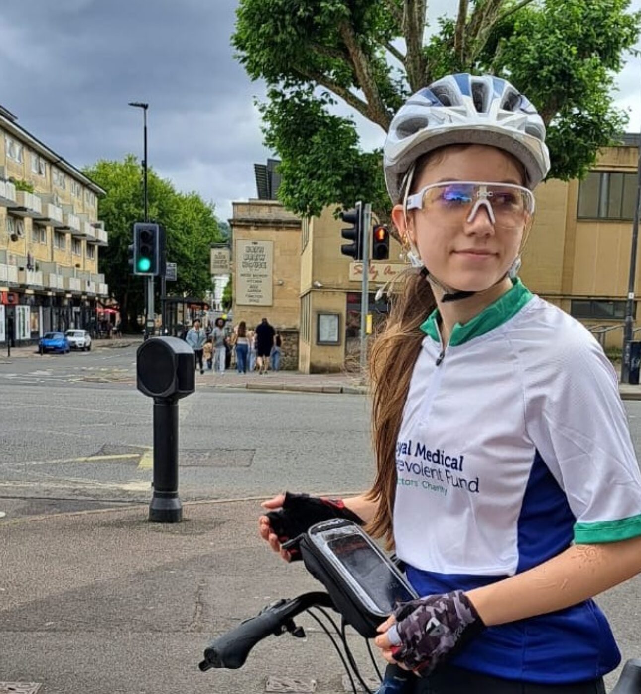 Isobel, stood on a street next to her bike, wearing a cylcing helmet and an RMBF cycling jersey