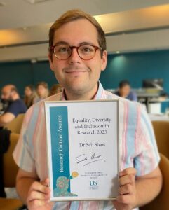 Portrait photo of Dr Sebastian Shaw. He is a young white man with brown hair and glasses. He's holding a framed award certificate that reads "Equality, Diversity and Inclusion in Research 2023".