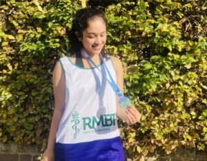 Ananya, standing outside with a hedge behind her, wearing an RMBF running vest, holding a medal and smiling