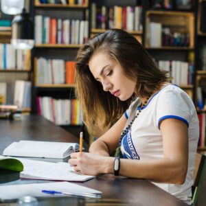 Female student sat at a desk, writing with a pencil, with a wall of books in the background.