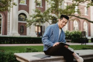 A young Asian man, sat on a plinth outside a University type building, smiling and looking at a book in his lap.