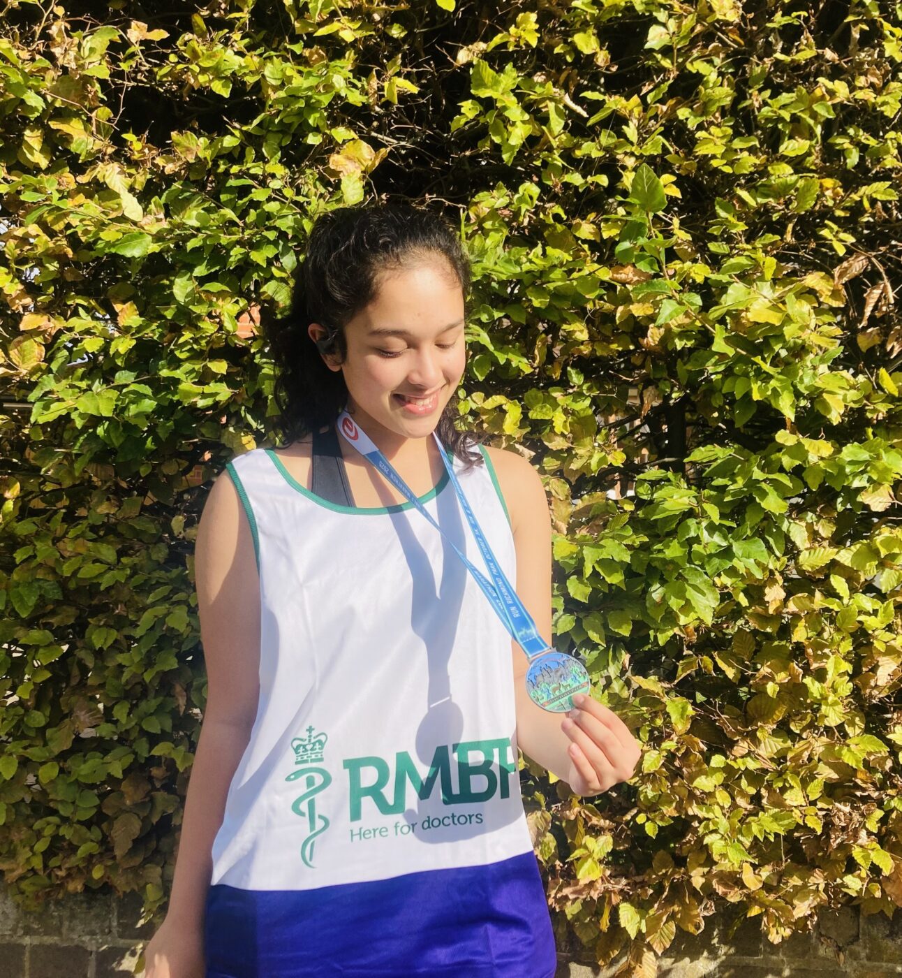 Student Ananya, standing in front of a hedge with sunlight. Ananya is smiling, wearing an RMBF running vest and looking at and holding a medal.