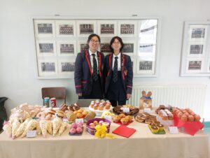Two female students, dressed in school uniform, smiling and standing behind a table filled with cakes