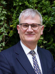 A photo of RMBF Trustee Dr John Holden. He is a white man with grey hair, wearing glasses and a suit and tie.