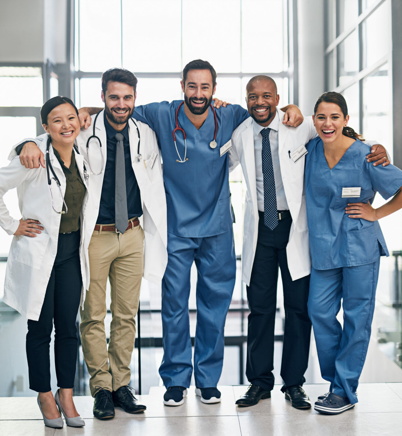 Group of 5 male and female doctors dressed in scrubs or white coat, smiling with their arms around each other.